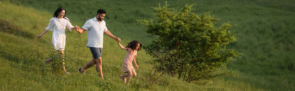 joyful family holding hands and running on green slope in countryside, banner.