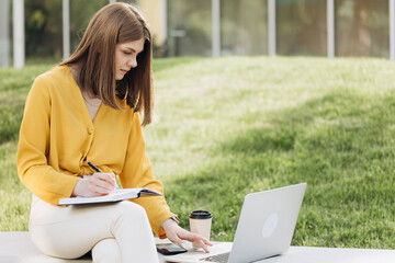 Portrait of focused student girl doing homework next to laptop computer. Teenage girl writing in notebook in front of laptop outdoors. Pretty girl studying at home education