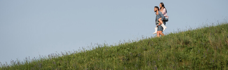 view from afar on man piggybacking woman in hilly meadow under blue sky, banner.