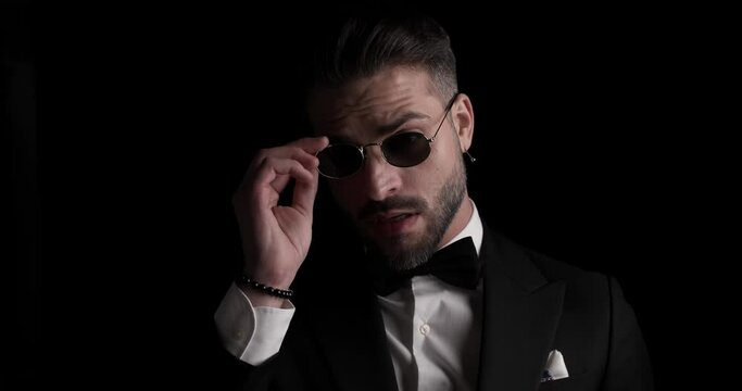 cool young groom in tuxedo arranging sunglasses, crossing arms and smiling in front of black background in studio
