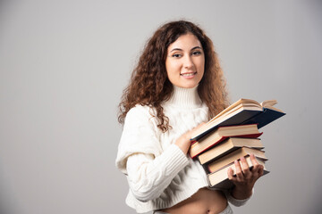 Young smiling woman carrying a lot of books on a gray wall