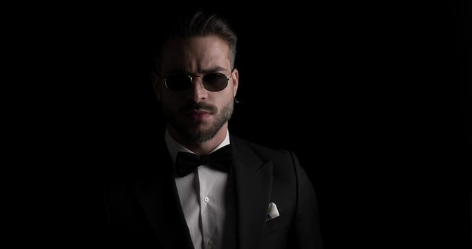 sexy groom with beard wearing sunglasses, adjusting tuxedo, crossing arms, looking to side and walking away in the dark