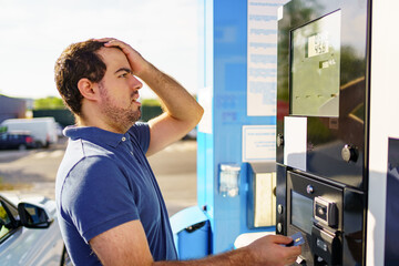 Caucasian young man surprised by high fuel prices on the gas station scoreboard.