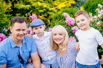 Fototapeta premium Portrait of family, caucasian husband his blond charming wife hugging with their cute serious emotions little son in garden with peonies on background.