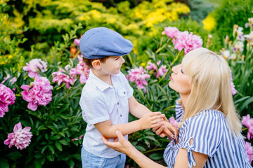 Motherhood and parenting. Relationship. Cute little lovely son and charming blond mother in home garden or park with peonies on background.