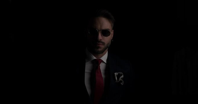 cool sexy businessman looking to side, adjusting tie and sunglasses and confidently folding arms on black background in the dark