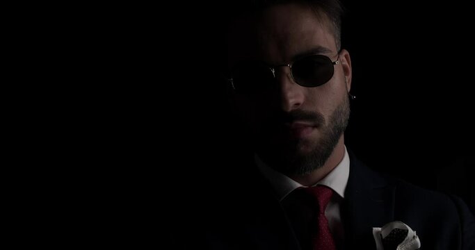 close up project video of unshaved man with sunglasses wearing suit looking to side and crossing arms on black background