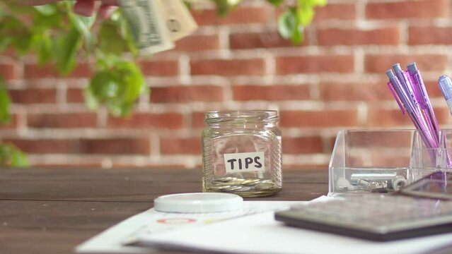 Tips are word written on sticker on jar lying on table. Hand puts money in nearest bank. Concept of tipping