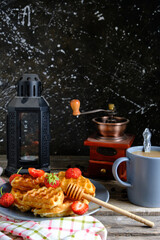 Waffles with strawberries and honey, sweet pastries plate and coffee mug, against background coffee grinder and kerosene lamp.