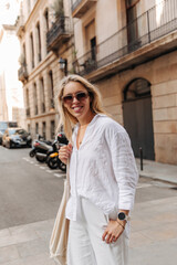 Fototapeta premium Young pretty woman staying on the street in sunglasses . European blonde smiling at camera wearing white linen suit 
