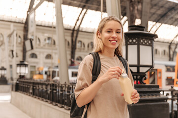 Beautiful european blonde woman staying with backpack wearing brown t-shirt . Smiling fair-haired lady holding in hands yellow bottle of water 