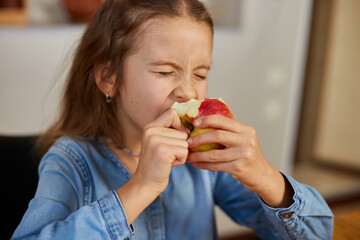 Cute little girl eat red apple at home at kitchen interior