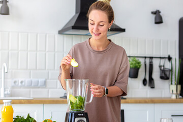 Young happy woman cooking healthy food. Caucasian female preparing breakfast from fresh green smoothie standing in the kitchen. Vegetarian, vegan food concept