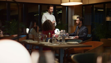 Waiter serving multiethnic couple on fancy restaurant dinner date at night.
