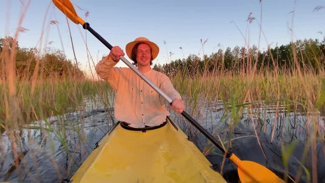 Young man sails on kayak through river bulrush at sunset. Guy smiles looking around with satisfied expression enjoying vacation in wild nature