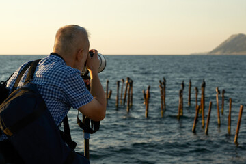 An adult man photographs a seascape with a flock of birds at sunset with a long-focus lens. The...