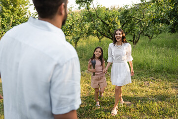 Fototapeta premium blurred man near happy wife and daughter holding hands near green trees.