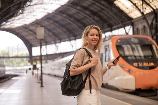 Picture of caucasian smiling woman looking at camera with phone . Focused on blonde staying on railway station with backpack