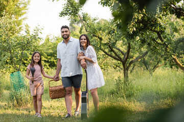 Fototapeta premium joyful family with wicker basket and guitar looking at camera in countryside garden.
