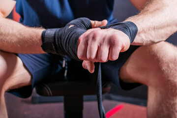 Cropped body of strong young mixed martial artist who sits on couch and wraps elastic bandage to fix his wrist for open fist fight.