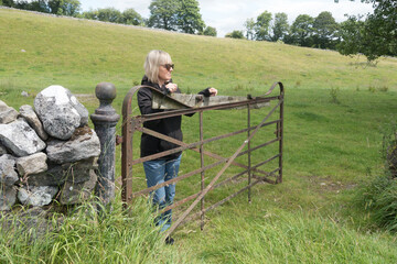An attractive blonde lady leans and rests on an old iron farm gate in rural setting with traditional dry stone wall.She wears sunglasses and is smiling.Yorkshire