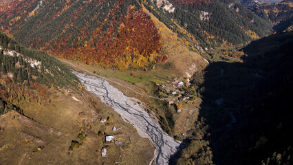 Fototapeta na wymiar Aerial view of a charming small village located down in the valley near forested hills. Footage. Flying over the mountains covered by colorful autumn trees.