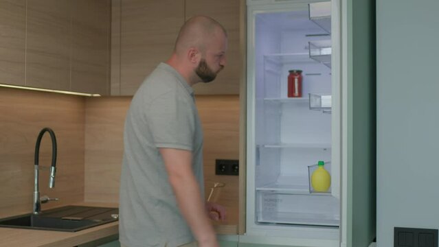 Hungry 30s man opening empty fridge and getting upset. Frustrated man wants to eat, but there is no food at home.