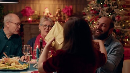 Excited husband giving his wife present while enjoying Christmas dinner with relatives. Happy man offering gift to young woman while celebrating winter holiday at home with loved ones. Handheld shot - Powered by Adobe