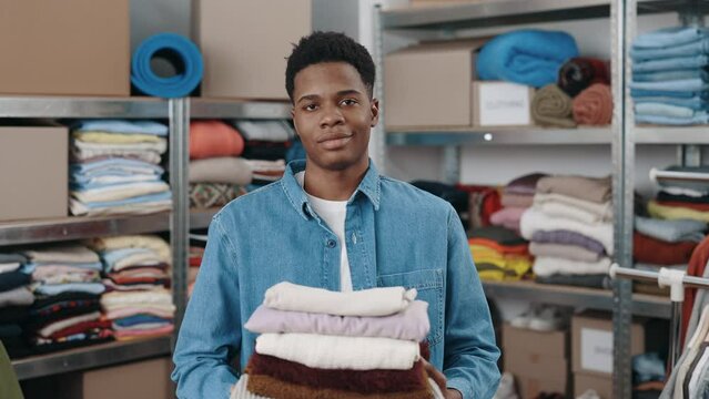 Portrait view of the multiracial male worker looking at the camera and stretching to the camera stack of clothes while working at the donating center or second hand. Humanitarian aid concept