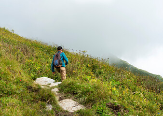 Naklejka na ściany i meble Rear view young man with a backpack walking uphill on a mountain path in fog among lush vegetation healthy and active lifestyle hiking outdoor activities, landscape