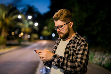 Bearded man wearing eyeglasses is holding scrolling texting in his cellphone at night street. Guy calls for taxi in an app in evening city - copy space and place for advertising