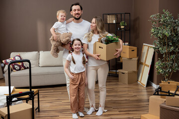 Fototapeta na wymiar Couple in love moves into a new apartment with their children, they bring cardboard boxes with packed things into the living room, a woman sets up decorations, they decorate their first home together.