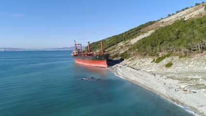Obraz premium Aerial for an empty industrial ship moored near sea shore with many people walking on a beach. Maritime cargo vessel standing near green trees slope in a summer sunny day.