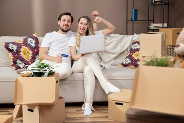 Portrait of a laughing young couple shopping online, sitting on a couch with a credit card and...