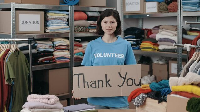 Portrait view of the caucasian volunteer woman holding banner with thank you phrase and looking at the camera with smile. Shelves with belongings at the background. Donations concept