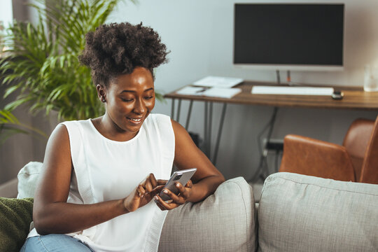 Shot of a young woman using a smartphone on the sofa at home. Smiling woman using mobile phone while sitting on sofa. Young female is text messaging in living room. She is relaxing at home.