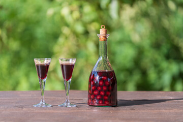 Homemade cherry brandy in two glasses and in a glass bottle on a wooden table in a summer garden