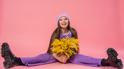 Portrait of a cute teenage girl with long hair in lilac fashionable clothes. a child holds a bouquet of yellow mimosa flowers, sitting full-length on a pink background in the studio.