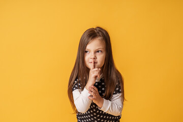portrait of a cute little girl pressing her index finger to her mouth stands on a yellow background. The child doesn't want to talk. Keeps a secret, learned a secret