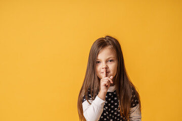 portrait of a cute little girl pressing her index finger to her mouth stands on a yellow...