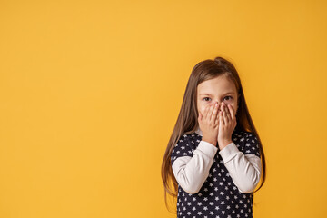 portrait of a cute little girl covering her mouth with both hands stands on yellow background. The...