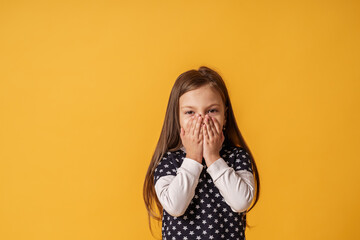 portrait of a cute little girl covering her mouth with both hands stands on yellow background. The child doesn't want to talk. Keeps a secret, learned a secret