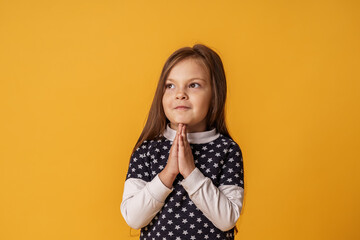 frustrated little girl, full hope, praying for something, child praying to God with his hands folded together, apologizing for bad behavior, looking away with pleading eyes.on yellow background