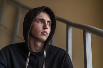 upset tense young man in a hood, sitting on the stairs and looking away, experiencing problems and...