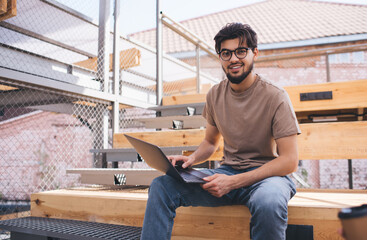 Male student sitting outdoors at university campus with laptop computer studying online. Young man freelancer sitting with netbook and looking at camera