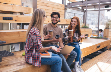 Three casual dressed happy friends talking and drinking take away coffee from cup while sitting outdoors. Group of smiling students recreating at campus having conversation. Spending time together