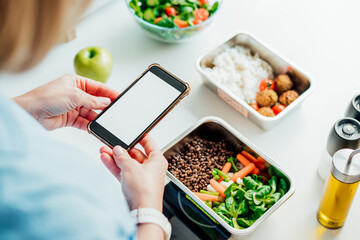 Fototapeta na wymiar Healthy diet plan for weight loss, daily ready meal menu. Woman using phone with blank screen for app while weighing lunch box cooked in advance on kitchen scale. Balanced portion. Pre-cooking