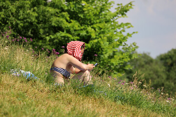 Girl in swimsuit sitting on the grass in summer park with smartphone in hands. Sunbathing on a nature, hot weather in city