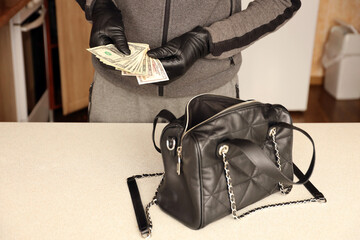 Robber in black outfit and gloves see in opened stolen women bag. The thief takes out US dollar...