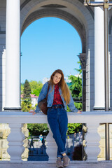 A woman in a casual denim outfit stands on a white open balcony against the backdrop of an arch and columns. Walk around the city sights. Architecture in classical style